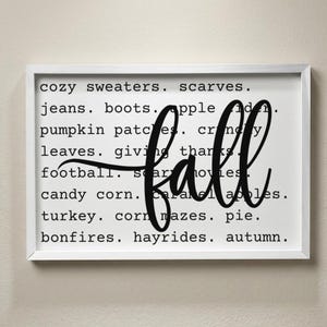May include: A rectangular wall sign in a white frame featuring a white background with black text listing fall-themed words including cozy sweaters, scarves, jeans, boots, apple cider, pumpkin patches, crunchy leaves, giving thanks, football, scary movies, candy corn, caramel apples, turkey, corn mazes, pie, bonfires, hayrides, and autumn, with the word fall written in large black script across the center.