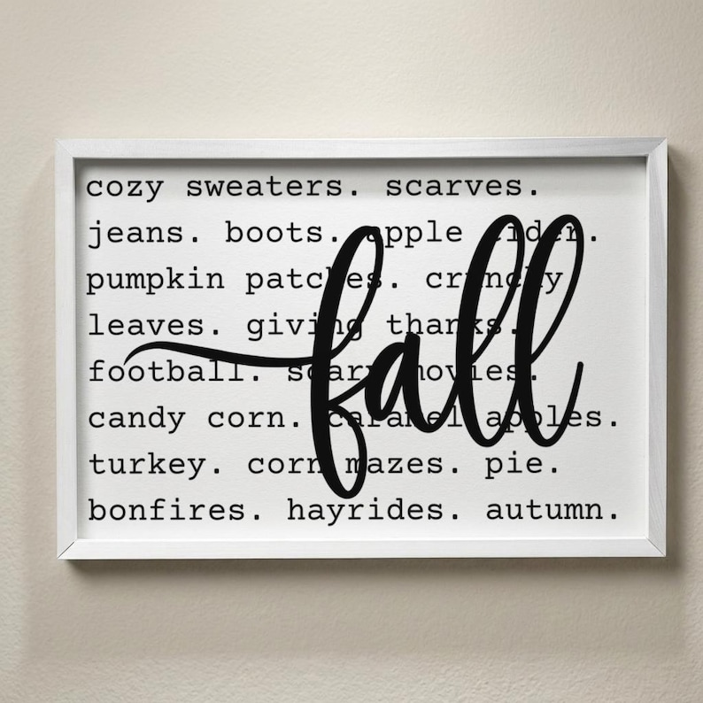 May include: A rectangular wall sign in a white frame featuring a white background with black text listing fall-themed words including cozy sweaters, scarves, jeans, boots, apple cider, pumpkin patches, crunchy leaves, giving thanks, football, scary movies, candy corn, caramel apples, turkey, corn mazes, pie, bonfires, hayrides, and autumn, with the word fall written in large black script across the center.