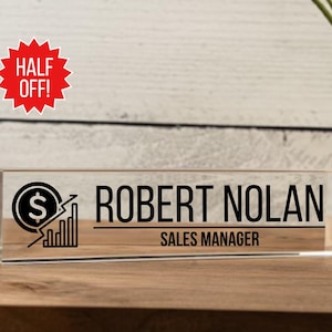 May include: A clear acrylic desk nameplate with a black graphic of a dollar sign and a rising bar chart. The nameplate reads "Robert Nolan, Sales Manager".