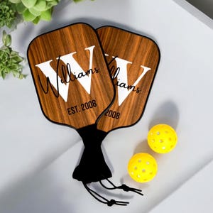 May include: Two wooden pickleball paddles with black handles and a black carrying bag. The paddles are personalized with the last name "Williams" and the year "EST. 2008".
