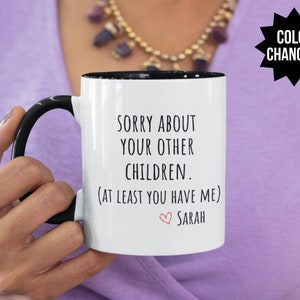 May include: White ceramic mug with a black rim and handle. The mug has black text that reads "Sorry about your other children. (At least you have me) Sarah" with a pink heart before the name.