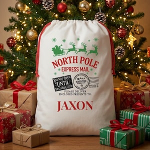 May include: A white drawstring gift sack with red cords featuring a green graphic of Santa in a sleigh with reindeer. Text reads: NORTH POLE EXPRESS MAIL, DO NOT OPEN UNTIL DECEMBER 25TH, and JAXON. It includes a North Pole postmark stamp.