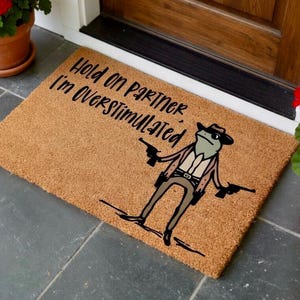 May include: A rectangular doormat made of natural fibers. The mat features a cartoon frog in a cowboy outfit holding two guns. The text "Hold on Partner I'm Overstimulated" is printed above the frog in black.