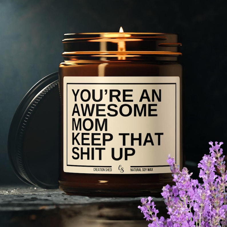 May include: A brown glass candle jar with a black label that reads "You're an Awesome Mom Keep That Shit Up". The candle is surrounded by lavender flowers.