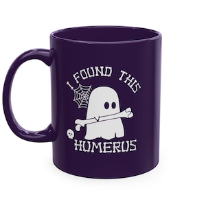 Op de afbeelding: Een donkerpaarse keramische mok met een witte afbeelding van een geest die een bot in de mond houdt. De tekst "I FOUND THIS HUMERUS" is in een botachtig lettertype boven en onder de geest gedrukt, vergezeld van een spinnenweb en een schedel.