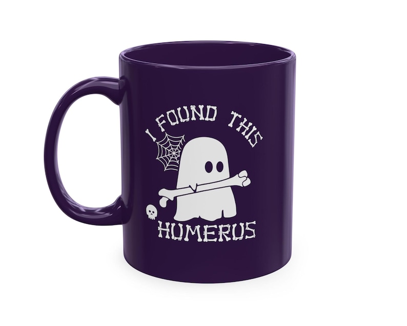 Op de afbeelding: Een donkerpaarse keramische mok met een witte afbeelding van een geest die een bot in de mond houdt. De tekst "I FOUND THIS HUMERUS" is in een botachtig lettertype boven en onder de geest gedrukt, vergezeld van een spinnenweb en een schedel.