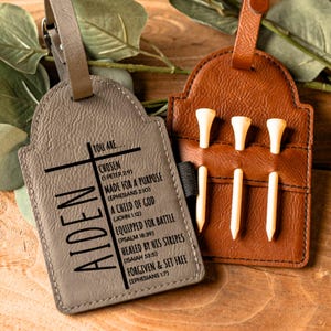 May include: A gray leather luggage tag with the name Aiden and a cross with bible verses. The tag is next to a brown leather golf tee holder with four golf tees.