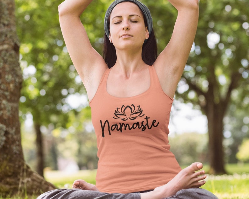May include: A woman wearing a peach tank top with a black lotus flower and the word "Namaste" printed on the front. She is sitting in a yoga pose with her eyes closed.