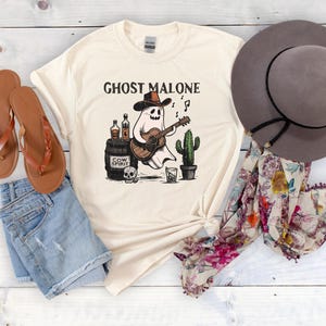 May include: A white t-shirt with a graphic of a ghost playing a guitar and holding a bottle of liquor. The text "Ghost Malone" is printed on the shirt. The shirt is laid out on a wooden surface with a pair of brown sandals, a brown hat, and a floral scarf.