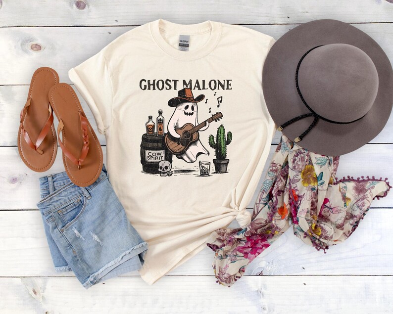 May include: A white t-shirt with a graphic of a ghost playing a guitar and holding a bottle of liquor. The text "Ghost Malone" is printed on the shirt. The shirt is laid out on a wooden surface with a pair of brown sandals, a brown hat, and a floral scarf.
