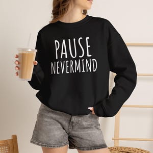 May include: A black long-sleeve sweatshirt featuring the white text PAUSE NEVERMIND printed on the front. The garment has a crew neckline and long sleeves, paired with gray denim shorts. The design is simple, featuring bold, sans-serif lettering.