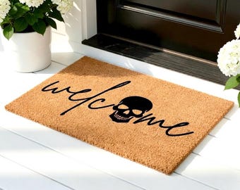 May include: A rectangular coir doormat with a brown background. The word "welcome" is written in a black cursive font, with a black skull graphic replacing the "o". The mat is placed in front of a black door.