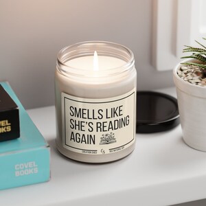 May include: A white candle in a clear glass jar with a black label that reads "Smells Like She's Reading Again". The label also includes the text "Creation Shed" and "9oz Natural Soy".