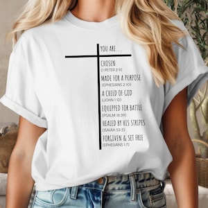 May include: White t-shirt with a black cross and text that reads "You are... Chosen (1 Peter 2:9) Made for a Purpose (Ephesians 2:10) A Child of God (John 1:12) Equipped for Battle (Psalm 18:39) Healed by His Stripes (Isaiah 53:5) Forgiven & Set Free (Ephesians 1:7)"