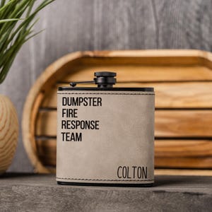 May include: A rectangular, light gray flask with black trim and a black cap. The text "DUMPSTER FIRE RESPONSE TEAM" and the name "COLTON" are printed on the front. The flask is displayed on a gray surface.