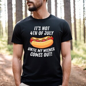 Pode incluir: Uma camiseta preta com um gr&aacute;fico branco de um cachorro-quente e o texto "N&atilde;o &eacute; 4 de julho at&eacute; que meu wiener saia!"