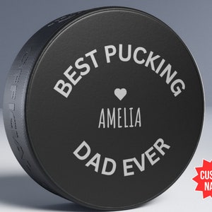 May include: Black hockey puck with white text that reads "Best Pucking Dad Ever" with a heart and the name "Amelia" in the centre.