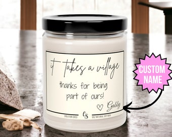 May include: A white candle jar with a black lid and a white label that reads "it takes a village thanks for being part of ours! Gabby". The label has a small heart symbol. The candle jar is on a gray countertop.