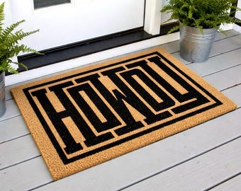 May include: A rectangular doormat with the word "HOWDY" in bold, black letters. The mat has a natural coir fiber background and a black border. The mat is placed on a gray wooden porch, near a white door and potted plants.