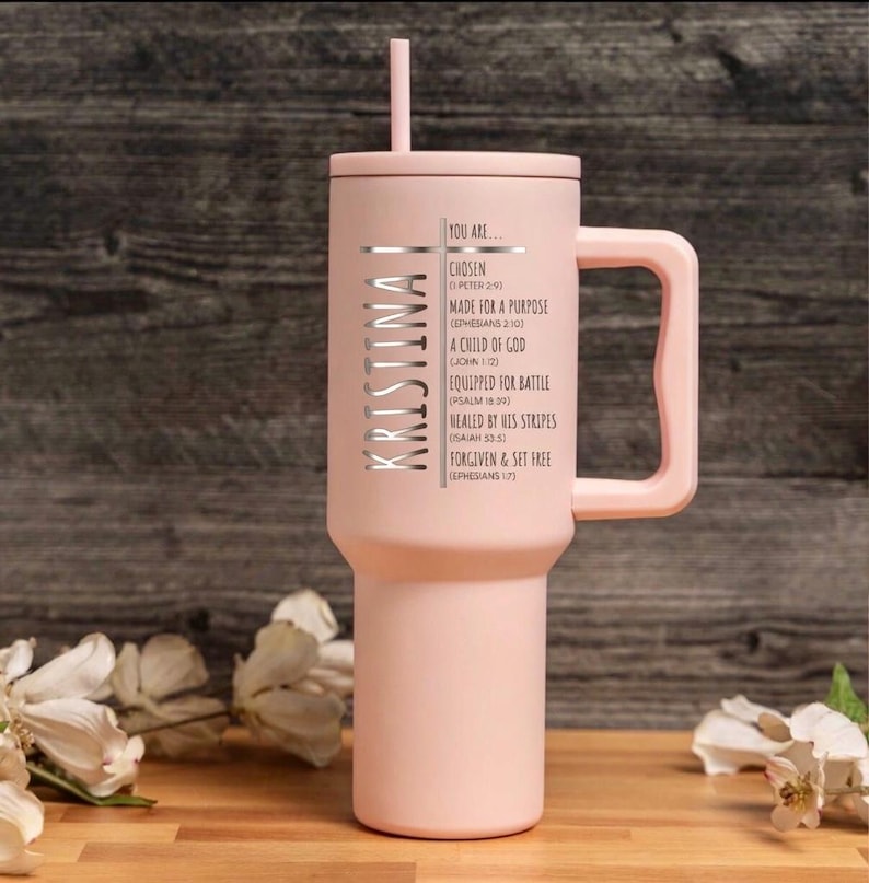 May include: A light pink tumbler with a handle and a straw. The name "KRISTINA" is vertically printed on the side, along with the words "YOU ARE..." and inspirational phrases. The tumbler is on a wooden surface.