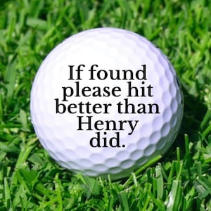 May include: A white golf ball with the text "If found please hit better than Henry did." The ball is resting on a bed of green grass.