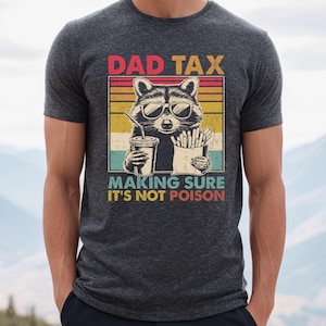 K&ouml;nnte beinhalten: Dunkelgraues T-Shirt mit einer Grafik eines Waschb&auml;ren mit Sonnenbrille, der ein Getr&auml;nk und Pommes h&auml;lt. Der Text lautet "DAD TAX MAKING SURE IT'S NOT POISON" im Retro-Stil.