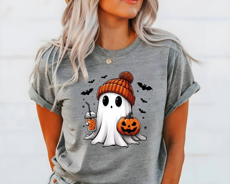 May include: Grey t-shirt featuring a cartoon ghost wearing a pumpkin-coloured hat and holding a pumpkin and a cup of orange juice. The ghost has black eyes and a small mouth. There are bats and stars around the ghost.