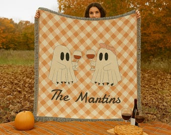 May include: A tan and white checkered throw blanket featuring two ghost illustrations holding wine glasses. The blanket has a fringed edge and the text "The Mantins" in a script font. A pumpkin and pie are in the foreground.