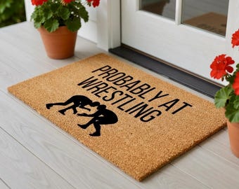 Op de afbeelding: Een rechthoekige deurmat van natuurlijke kokosvezel. De mat heeft de tekst "PROBABLY AT WRESTLING" in zwarte blokletters, met een silhouet van twee worstelaars. De mat ligt op een lichtgrijze houten ondergrond.