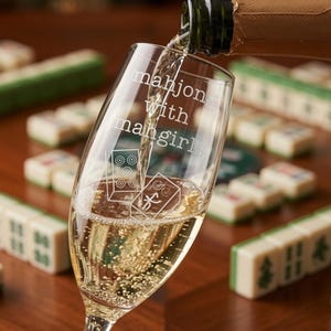 K&ouml;nnte beinhalten: Ein klares Sektglas aus Glas, das mit Sekt gef&uuml;llt wird. Das Glas ist mit dem Text "mahjong with mahgirls" und zwei Abbildungen von Mahjong-Steinen graviert. Das Glas ist transparent und zeigt deutlich sichtbare Bl&auml;schen.