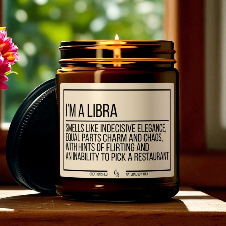 K&ouml;nnte beinhalten: Eine braune Glas-Kerze mit schwarzem Deckel. Die Kerze hat ein wei&szlig;es Etikett mit dem Text "I'M A LIBRA" und einer Beschreibung des Duftes. Die Kerze ist angez&uuml;ndet, und der Hintergrund ist unscharf.