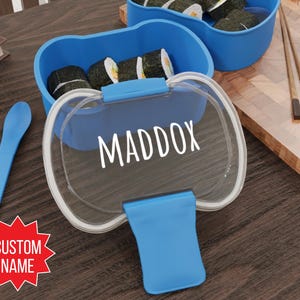 May include: Blue plastic lunchbox with a clear lid and a blue handle. The lunchbox has the name "MADDOX" printed on the lid. The lunchbox is sitting on a wooden table with a spoon and chopsticks.