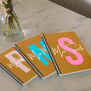Puede incluir: Tres cuadernos de espiral marrones con letras de colores en la portada. El primer cuaderno tiene una "P" rosa con "Sra." escrito en cursiva arriba. El segundo cuaderno tiene una "M" azul con "Sra." escrito en cursiva arriba. El tercer cuaderno tiene una "S" rosa con "Sra. Smith" escrito en cursiva arriba.