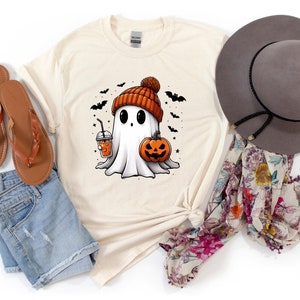 May include: A beige t-shirt featuring a cartoon ghost wearing an orange and black knitted hat and holding a pumpkin and a cup of orange juice. The ghost is surrounded by bats and stars.