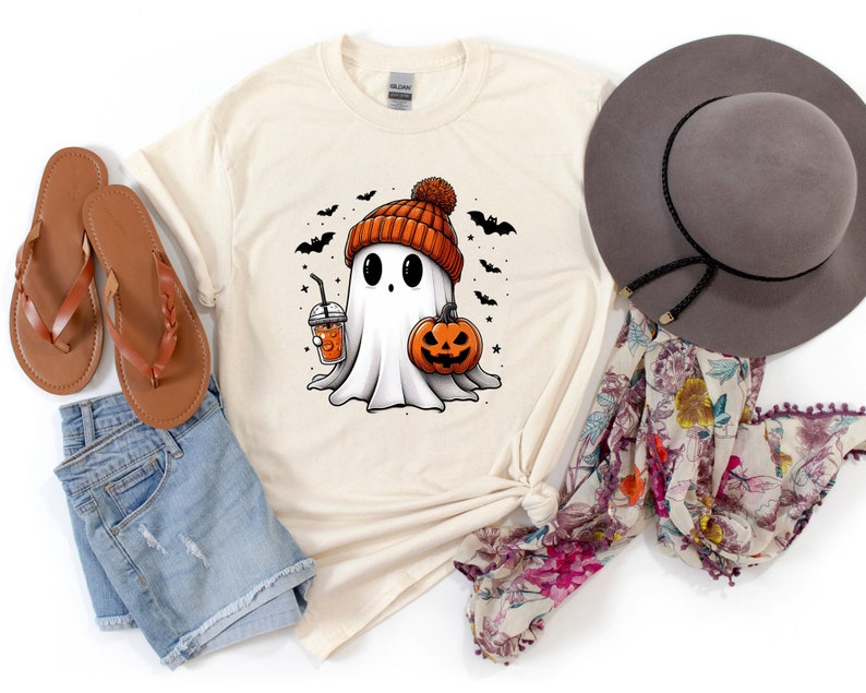 May include: A beige t-shirt featuring a cartoon ghost wearing an orange and black knitted hat and holding a pumpkin and a cup of orange juice. The ghost is surrounded by bats and stars.