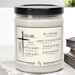 May include: A white candle with a black lid and a cream label. The label has a black cross and the text "GABRIEL YOU ARE... chosen (1 PETER 2:9) made for purpose (EPHESIANS 2:10) a child of God (JOHN 1:12) His workmanship (EPHESIANS 2:10) equipped with strength for the battle (PSALM 18:39) healed by His stripes (ISAIAH 53:5) forgiven and set free (EPHESIANS 1:7) CREATION SHED Cs 9OZ NATURAL SOY WAX".