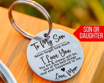 May include: A silver-colored keychain with a round pendant. The pendant has the text "To My Son, I Love You" and a message from "Mom". A red arrow indicates "Son or Daughter". The keychain is on a wooden surface.