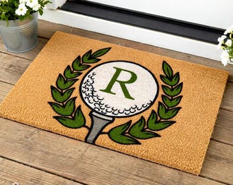 May include: A rectangular doormat with a golf theme. The mat is tan with a green and white golf ball design in the centre, featuring the letter "R". The design is surrounded by green laurel leaves.