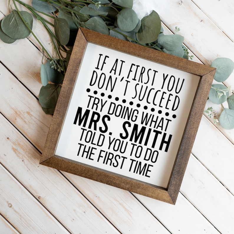 May include: A square wooden sign with a dark brown frame and a white background. The sign features black text that reads: IF AT FIRST YOU DON'T SUCCEED TRY DOING WHAT MRS. SMITH TOLD YOU TO DO THE FIRST TIME. The text is in a sans-serif font.