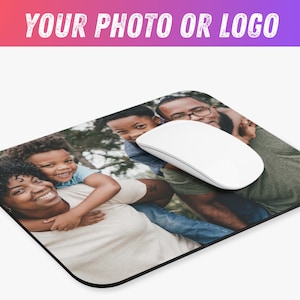 May include: A family of four, two adults and two children, are smiling and looking at the camera. The family is standing in a grassy area with trees in the background. The image is printed on a black mouse pad.