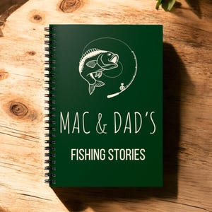 May include: A dark green spiral bound notebook with a white illustration of a fish jumping out of the water with a fishing rod. The text "MAC & DAD'S FISHING STORIES" is printed in white on the cover.