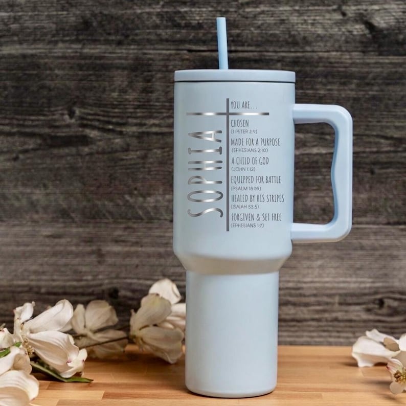 May include: A light blue insulated tumbler with a handle and a straw. The tumbler has the name "SOPHIA" in silver lettering on the side, along with inspirational text and a cross design. The tumbler is designed to hold cold beverages.