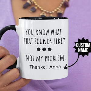 Könnte beinhalten: Weiße Keramiktasse mit schwarzem Henkel und Rand. Die Tasse hat schwarzen Text, der lautet "YOU KNOW WHAT THAT SOUNDS LIKE? ... NOT MY PROBLEM. Thanks! Anna" mit einem Pfeil, der nach rechts zeigt. Ein schwarzer Stern mit dem Text "CUSTOM NAME" befindet sich in der oberen rechten Ecke.