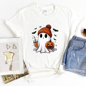 May include: White t-shirt featuring a cartoon ghost wearing a brown and orange knitted hat and holding a pumpkin and a cup of orange drink. The ghost has large eyes and a small mouth. There are bats flying around the ghost.