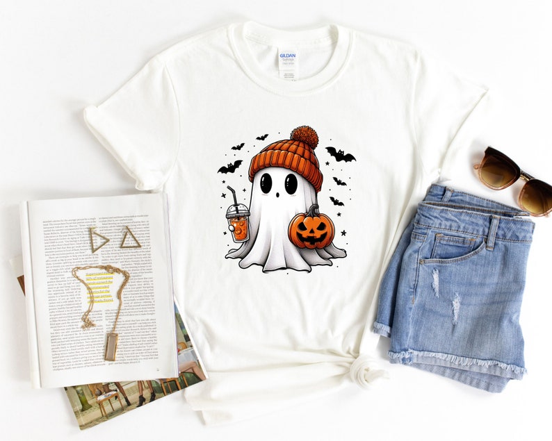 May include: White t-shirt featuring a cartoon ghost wearing a brown and orange knitted hat and holding a pumpkin and a cup of orange drink. The ghost has large eyes and a small mouth. There are bats flying around the ghost.