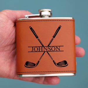 May include: A brown leather-wrapped flask with a silver top and a personalized inscription that reads "JOHNSON" with two crossed golf clubs above the name.