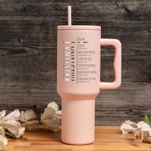 May include: A light pink tumbler with a handle and a straw. The name "KRISTINA" is vertically printed on the side, along with the words "YOU ARE..." and inspirational phrases. The tumbler is on a wooden surface.