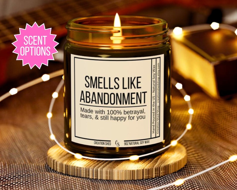 May include: A brown glass candle with a gold lid and a white label that reads "Smells Like Abandonment". The label also says "Made with 100% betrayal, tears, & still happy for you".