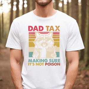 K&ouml;nnte beinhalten: Wei&szlig;es T-Shirt mit einer Grafik, die einen Waschb&auml;ren mit Sonnenbrille zeigt, der ein Getr&auml;nk und Pommes h&auml;lt. Der Text lautet "DAD TAX MAKING SURE IT'S NOT POISON" in einem Retro-Farbschema.