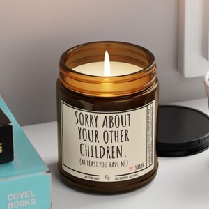 May include: A lit soy wax candle in a brown glass jar with a black lid. The candle has a white label with the text "SORRY ABOUT YOUR OTHER CHILDREN. (AT LEAST YOU HAVE ME)." The candle is next to a small potted plant and books.